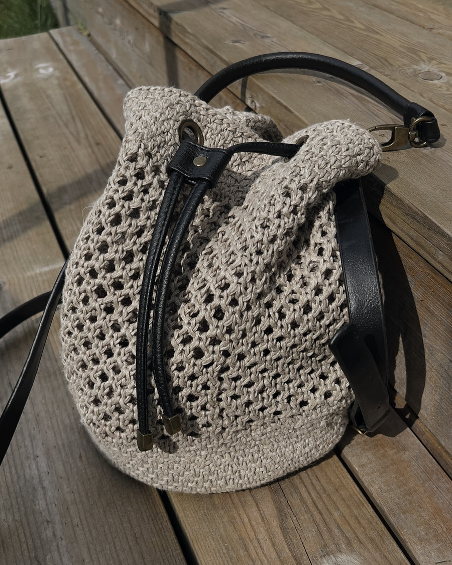Bucket bag i nätmönster i naturfärgat lin på trägolv, med svarta läderremmar · stickas utan sömmar